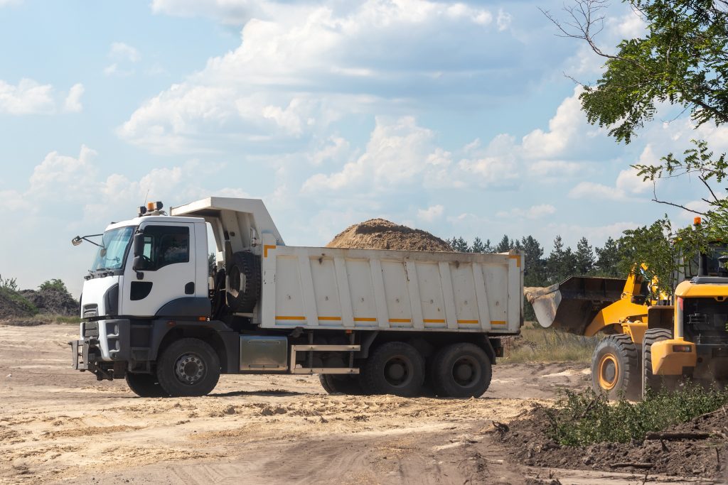 Dump truck with a bunch of land in the back and excavator at the construction site outside the city. Road works on an intercity highway on a cloudy day. Selective focus